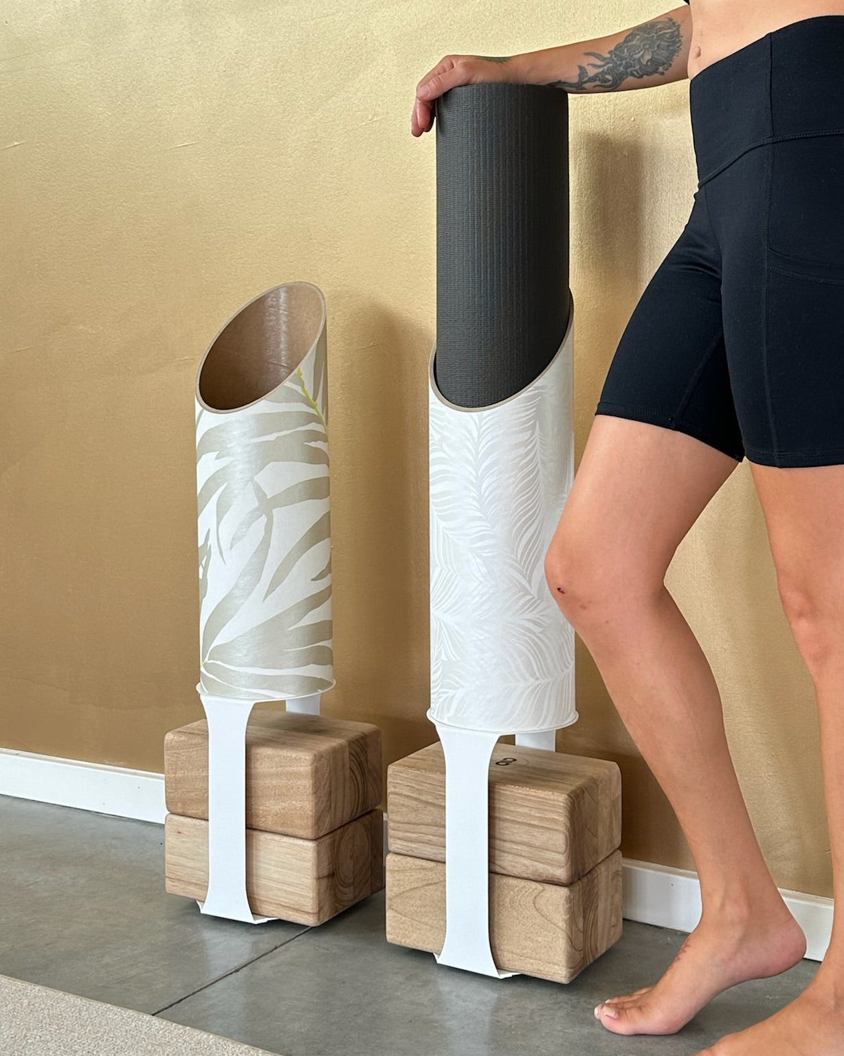 Person in black athletic wear standing next to yoga blocks and a mat against a gold wall.