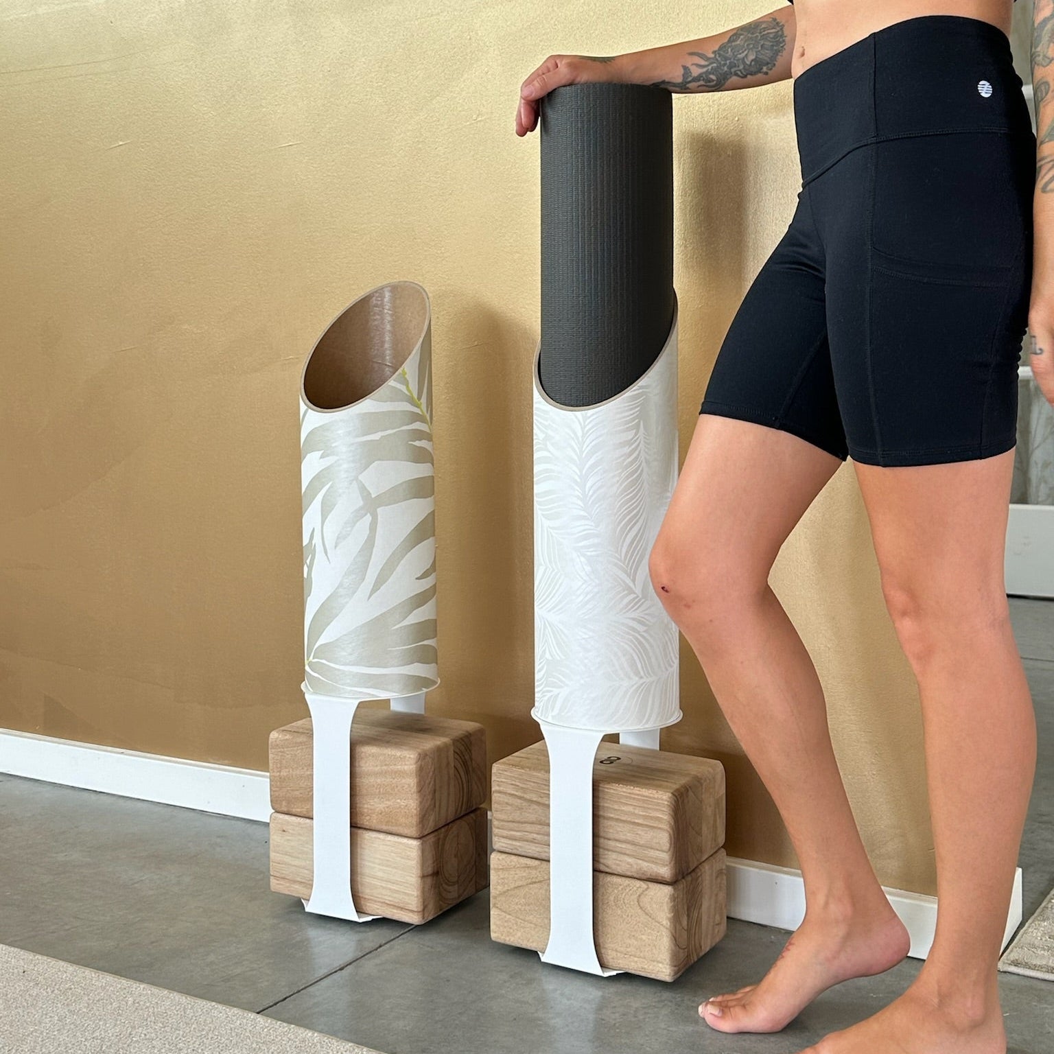 Person in black athletic wear standing next to yoga blocks and a mat against a gold wall.
