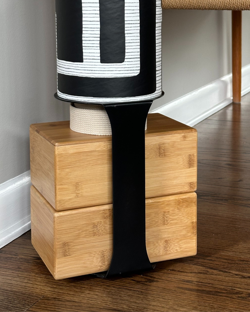 A yoga storage bundle consisting of a yoga mat tube with a geometric pattern, in black, gold, and white colors, resting on two stacked bamboo blocks.