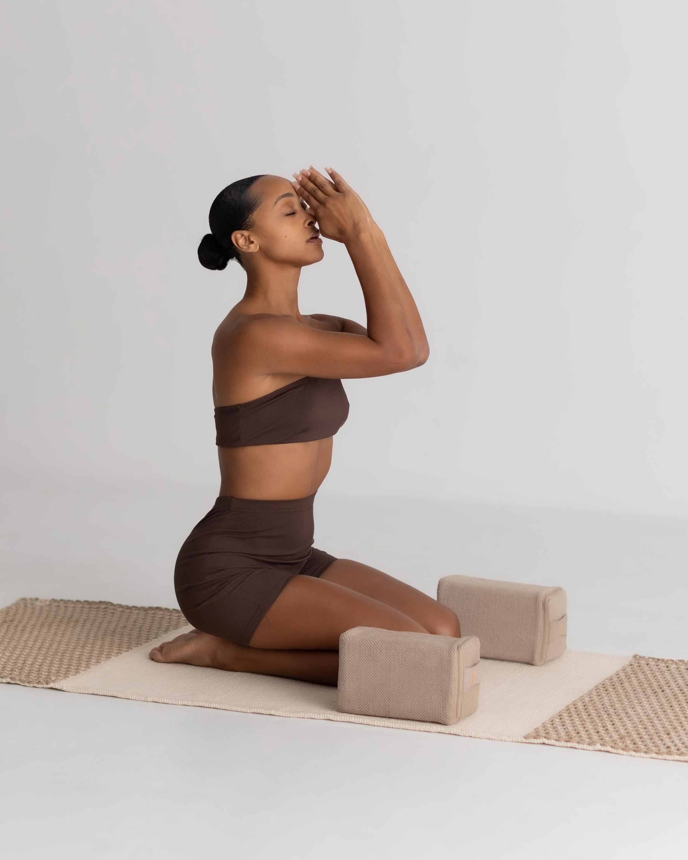 Woman in a brown outfit practicing yoga on a mat with blocks against a white background