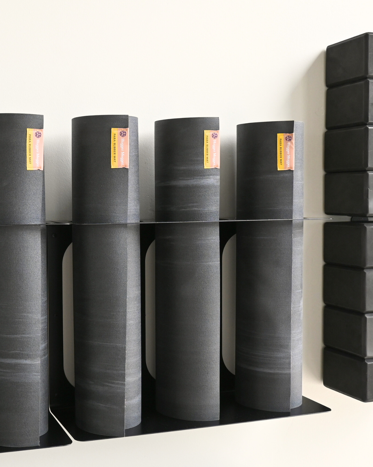 Several dark gray yoga mats are stacked on black metal shelves against a plain white wall, with a stack of black yoga blocks on the right side.