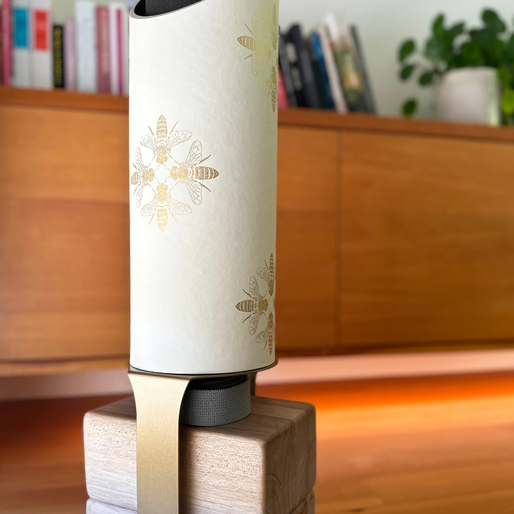 White cylindrical device with bee designs on a wooden stand, blurred books and plant in the background
