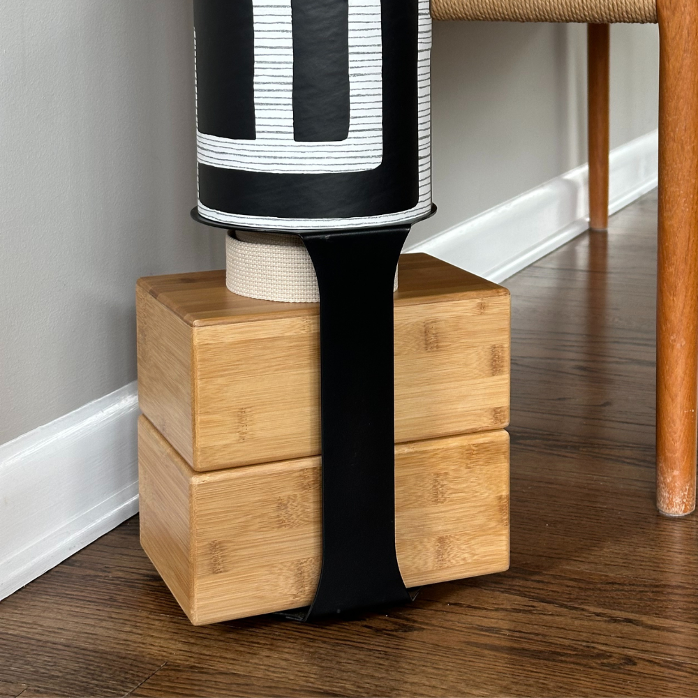 A yoga storage bundle consisting of a yoga mat tube with a geometric pattern, in black, gold, and white colors, resting on two stacked bamboo blocks.