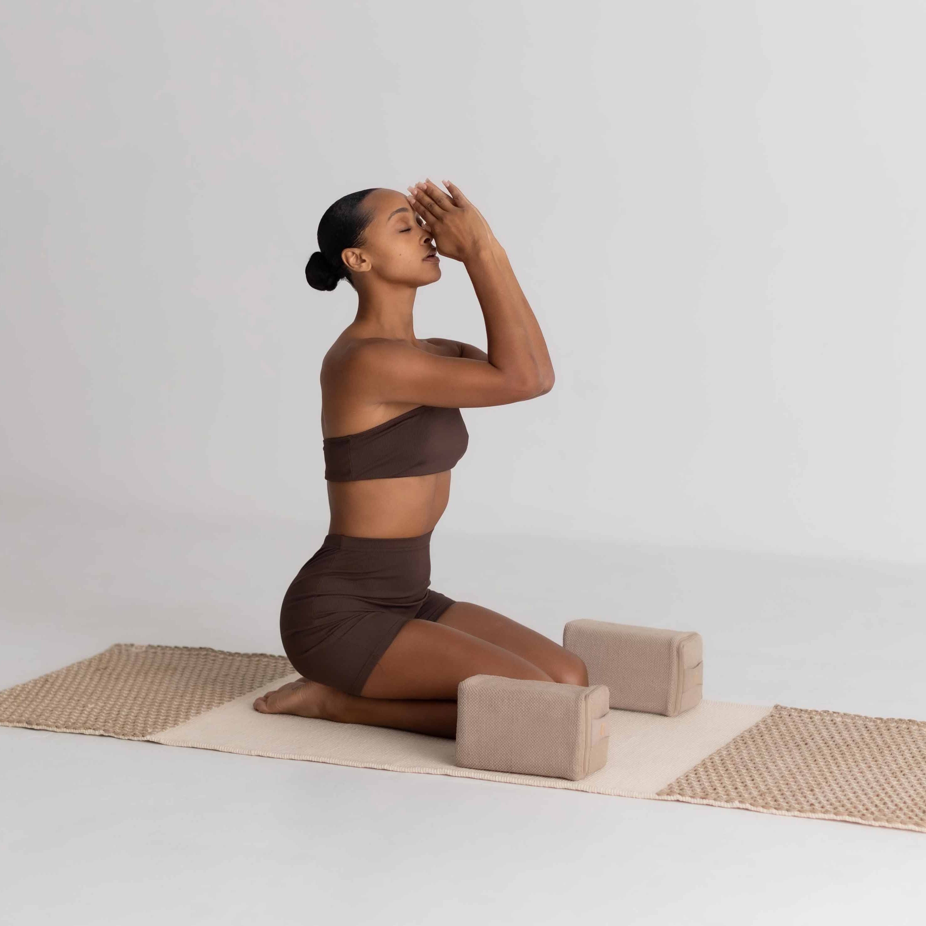 Woman in a brown outfit practicing yoga on a mat with blocks against a white background
