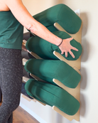 A person wearing a green tank top and dark patterned leggings is placing a dark green yoga bolster onto a light wood wall-mounted rack.