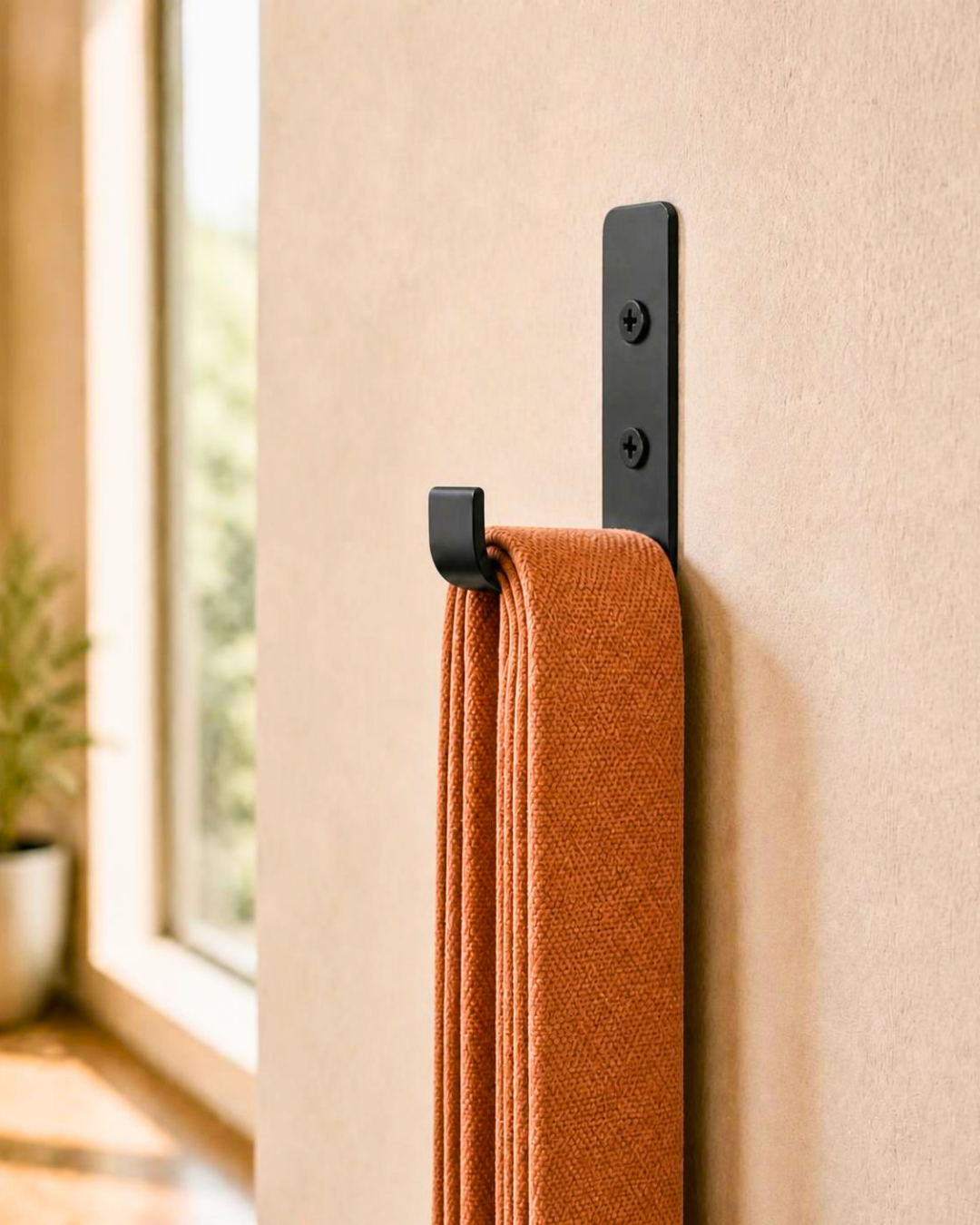 A close-up of a black wall-mounted hook holding a stack of folded terracotta orange yoga straps, set against a beige textured wall with a softly blurred window and plant in the background.