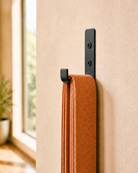 A close-up of a black wall-mounted hook holding a stack of folded terracotta orange yoga straps, set against a beige textured wall with a softly blurred window and plant in the background.