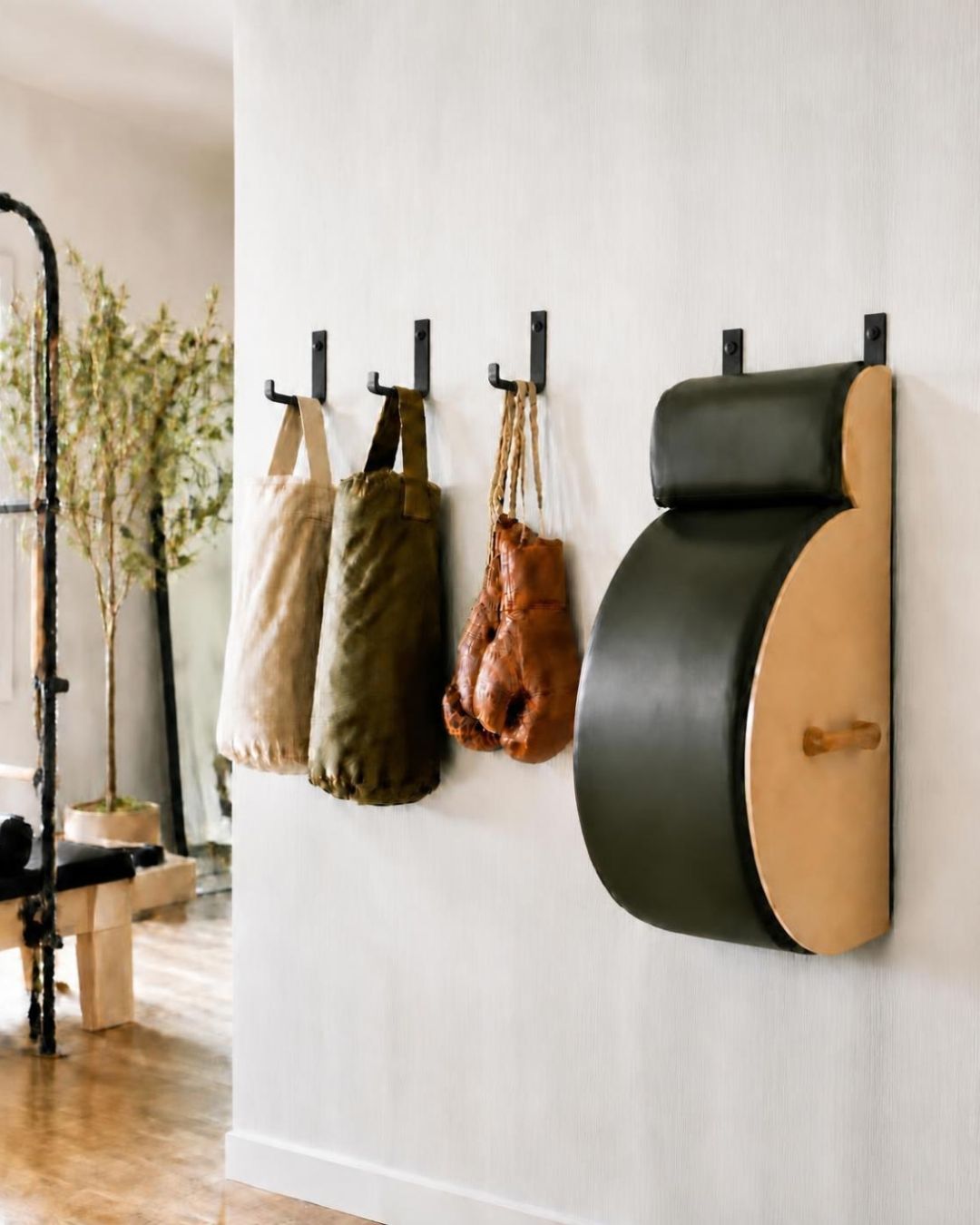 Wall-mounted fitness gear displayed on a white wall in a studio interior. The ensemble includes a spine corrector, used in pilates, on the right, and two hanging sand bags in beige, olive green, and a brown pair of boxing gloves, each attached to a black hook.