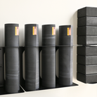 Several dark gray yoga mats are stacked on black metal shelves against a plain white wall, with a stack of black yoga blocks on the right side.