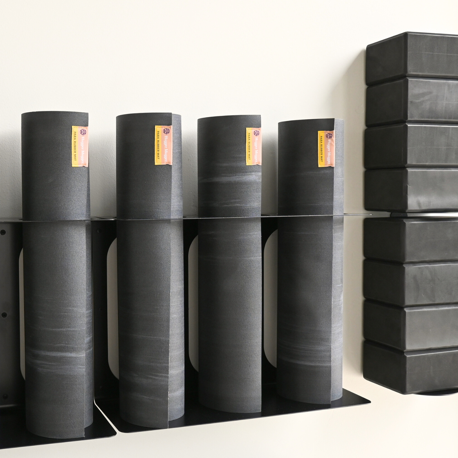 Several dark gray yoga mats are stacked on black metal shelves against a plain white wall, with a stack of black yoga blocks on the right side.